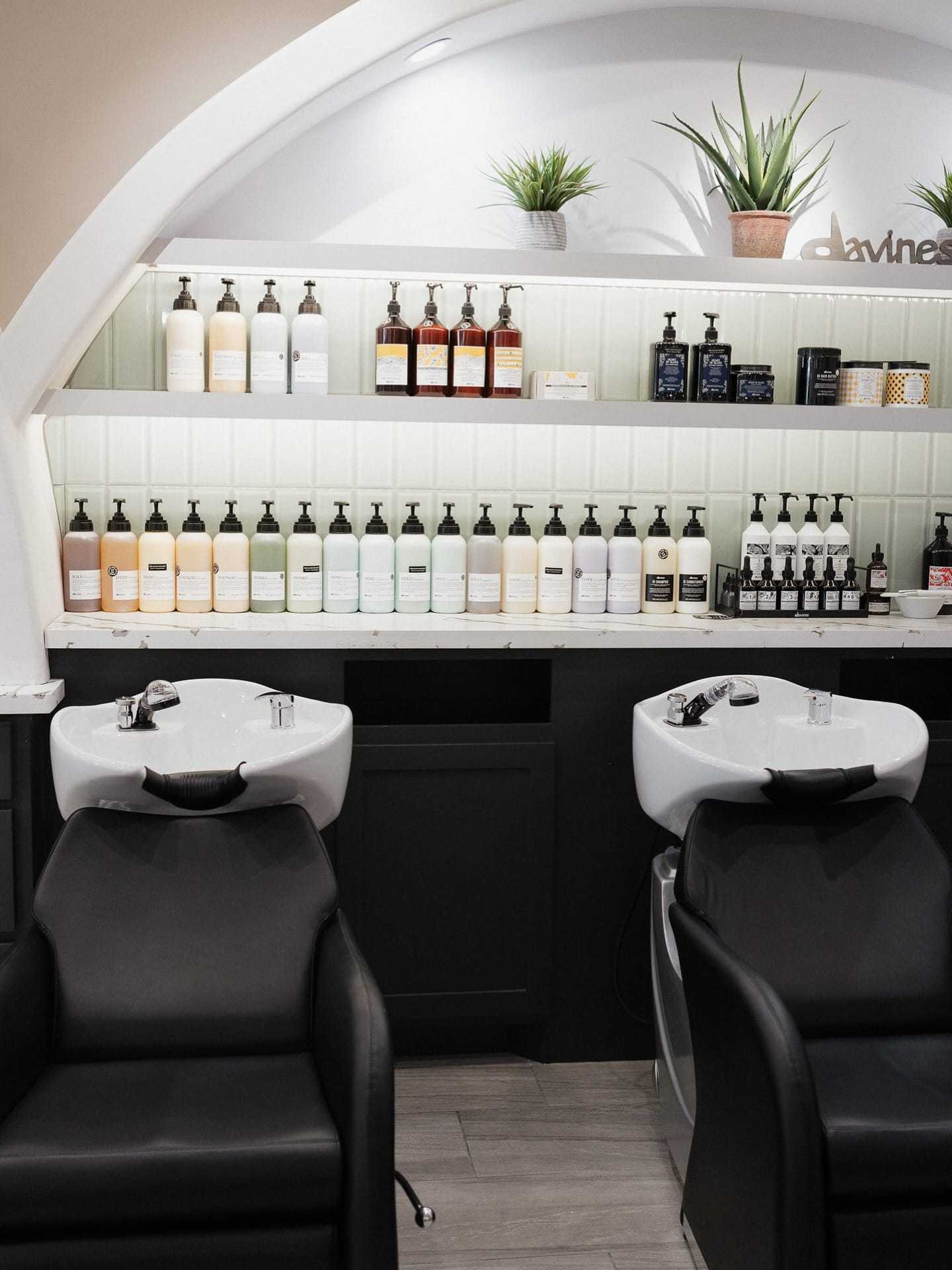 Hair salon with shampoo bottles on shelves and black wash basins in front.
